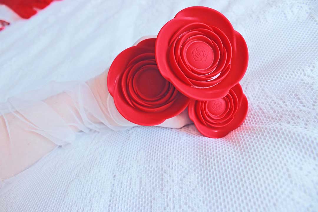 Gloved hand holding 3 red rose butt plugs on a white lace blanket