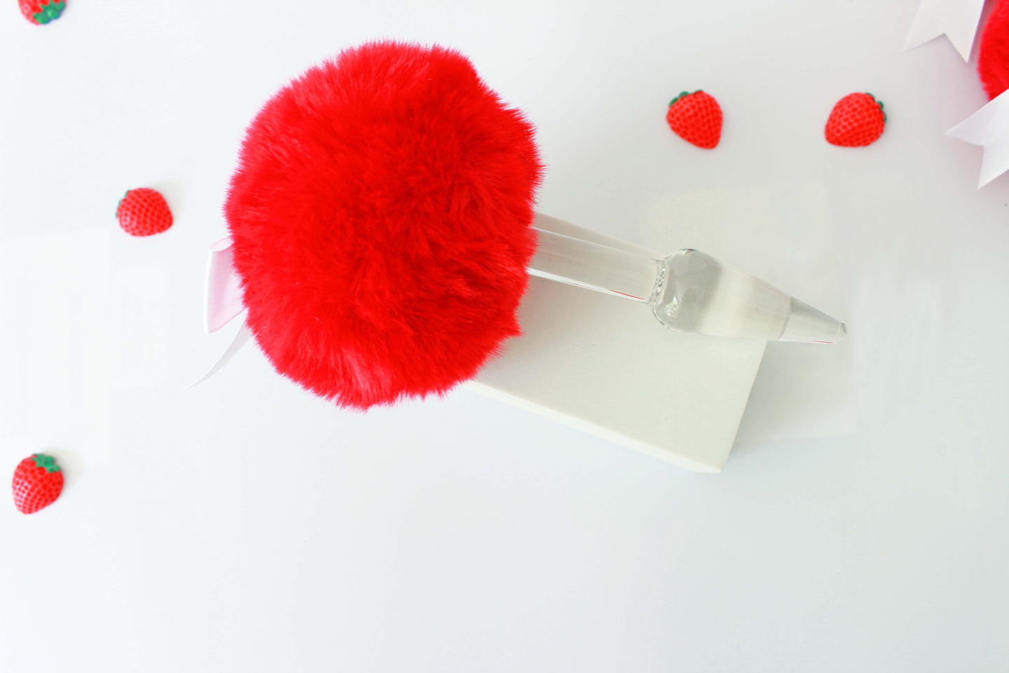 A red bunny tail butt plug with a white bow set on a stand showing off the glass plug portion on a white background with strawberries.