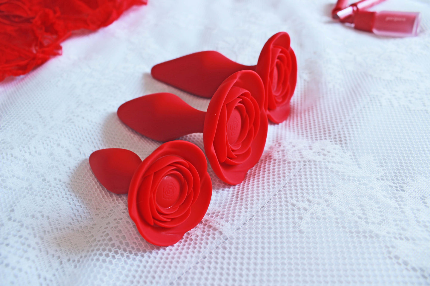 Small red rose butt plug laid in front of medium and large sizes of the same butt plug on white background with indistinguishable red objects in background