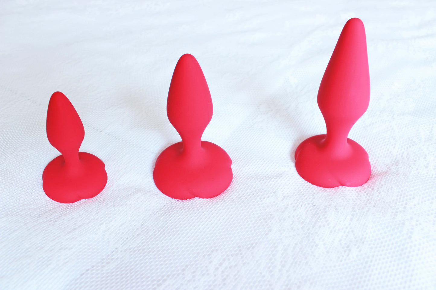 3 red rose silicone anal training butt plugs standing flat on their bases on a white lace blanket