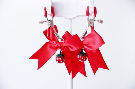 Silver adjustable tension screw nipple clamps with red pvc caps and pred bows with red strawberry bells hanging from them on a jewlery stand on a white background.