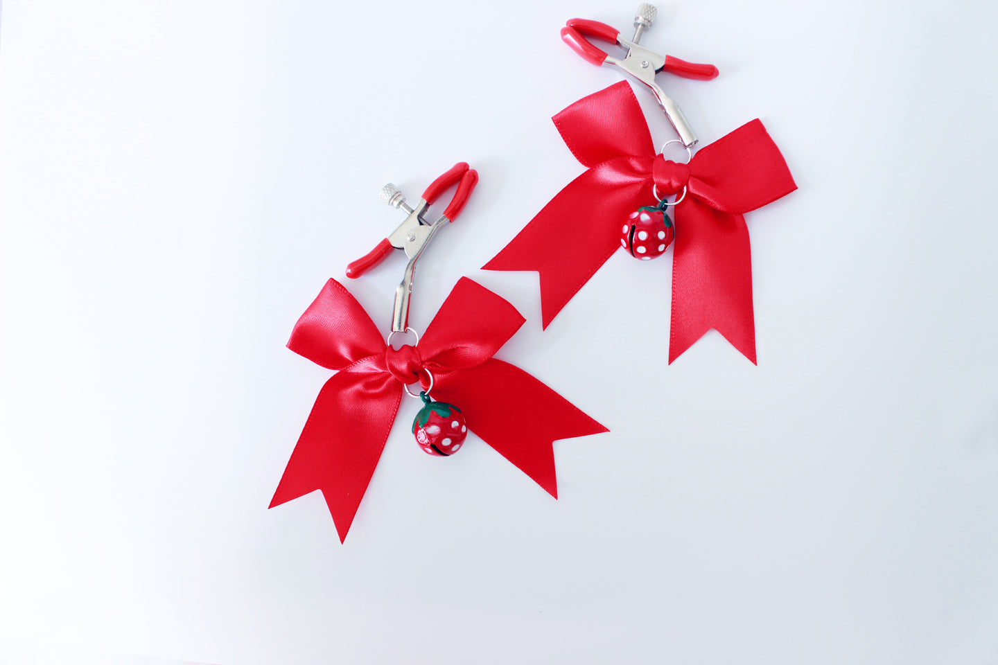 Silver ajustable screw nipple clamps with red pvc cap ends and red strawberry bells with red bows hanging from them on a white background.