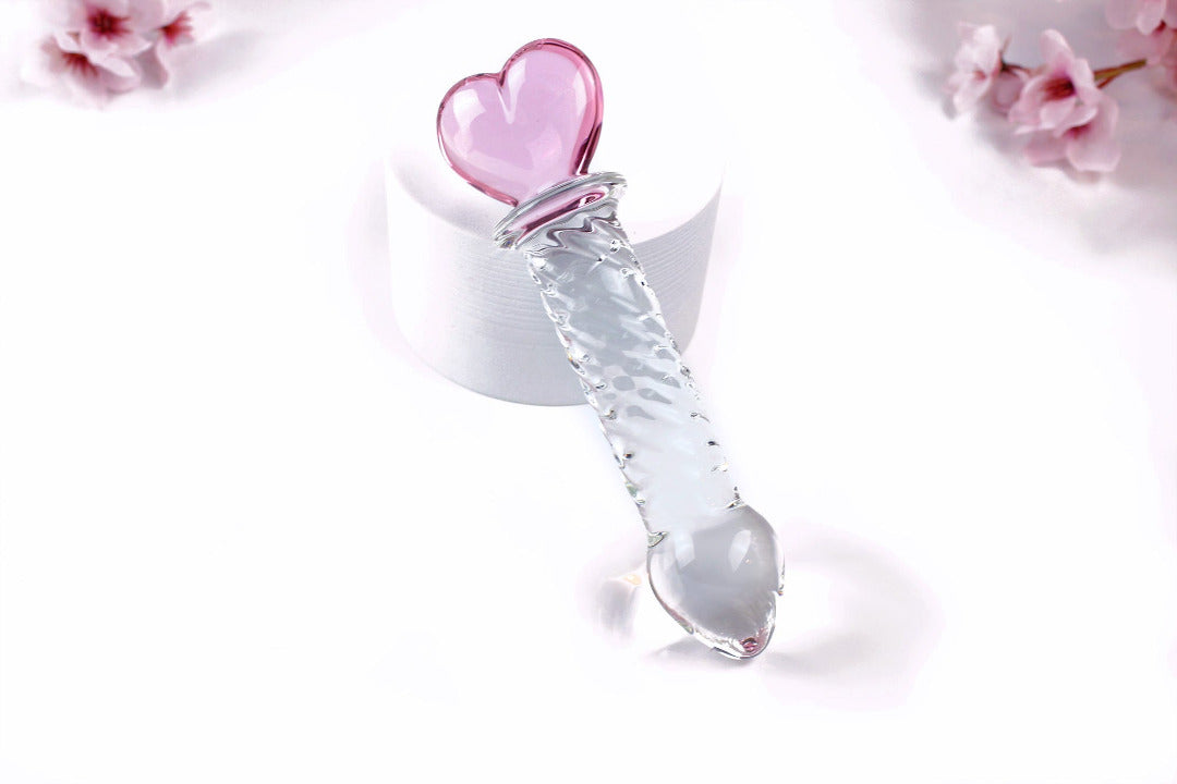 Glass ridged dildo with a clear shaft and a pink heart base on a white cylinder elevated on a white background with flowers