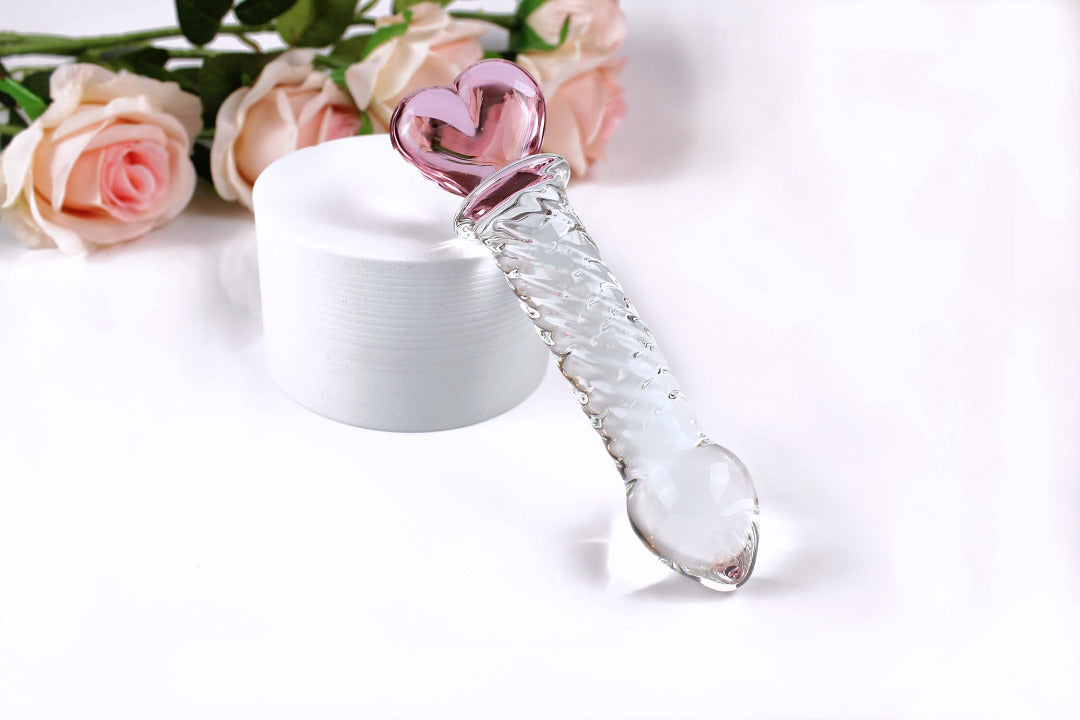 Glass ridged dildo with a clear shaft and a pink heart base on a white cylinder elevated on a white background with flowers