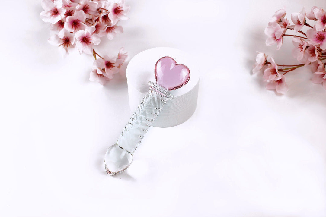 Glass ridged dildo with a clear shaft and a pink heart base on a white cylinder elevated on a white background with flowers