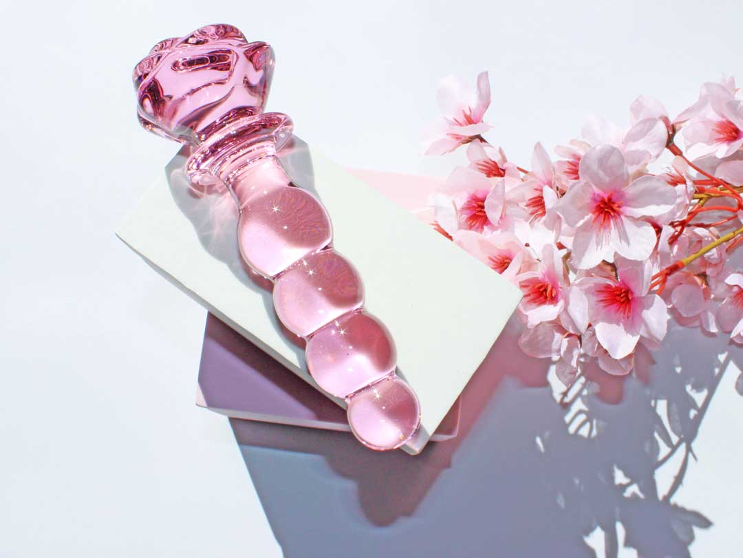 Pink glass dildo with a rose base and a knotted shaft elevated on a white square against a white backdrop with flowers.