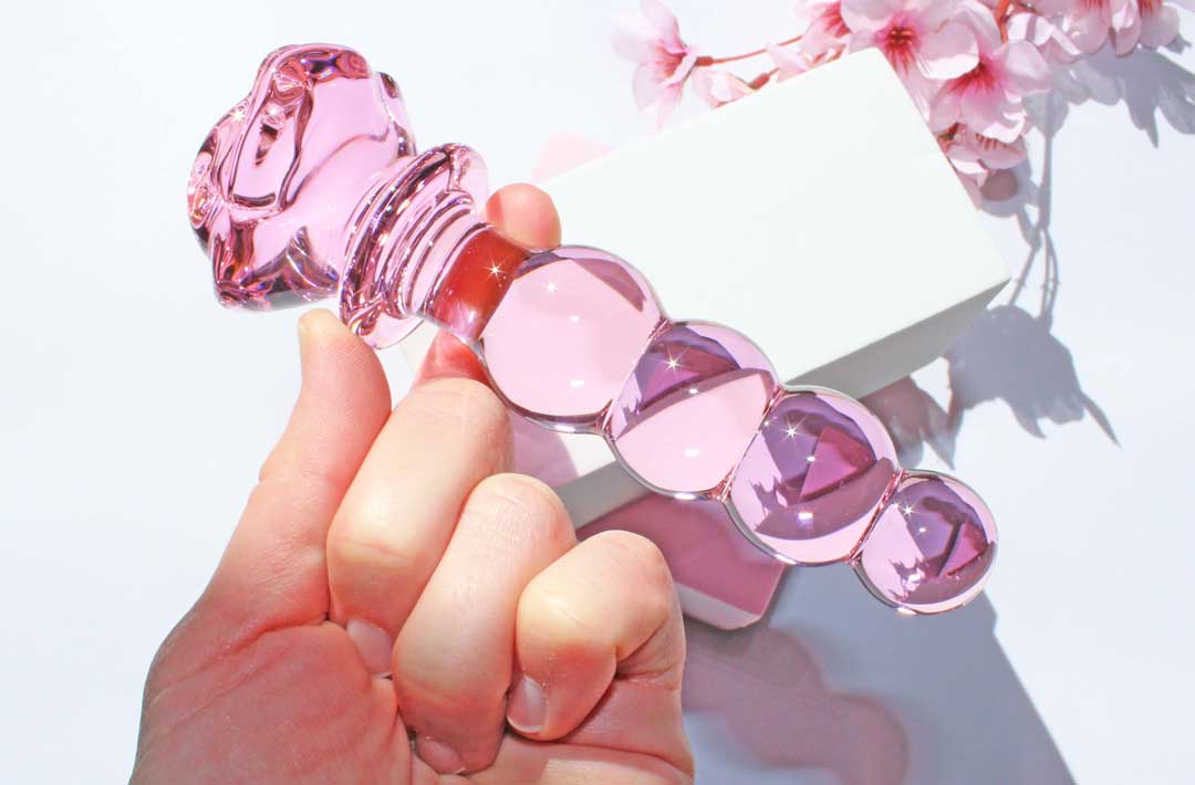 Pink glass dildo with a rose base and a knotted shaft being held over a white backdrop with flowers.