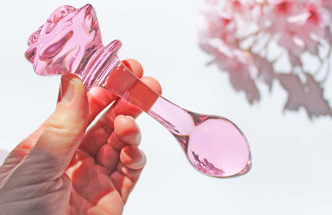 Pink glass butt plug with a rose base held above a white backdrop with flowers.