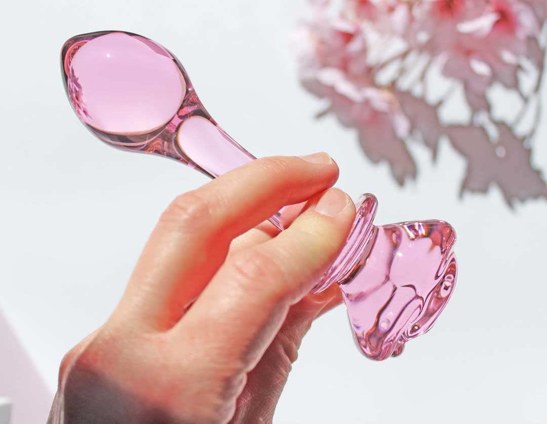 Pink glass butt plug with a rose base held above a white backdrop with flowers.