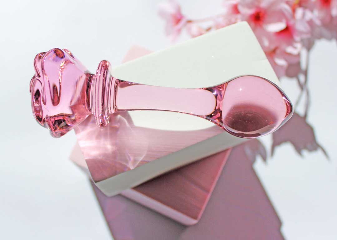 Pink glass butt plug with a rose base elevated on a white square against a white backdrop with flowers.