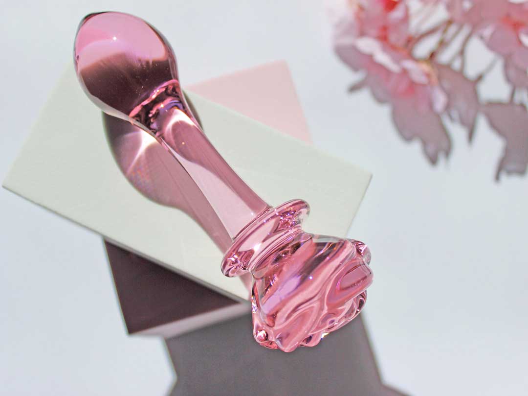 Pink glass butt plug with a rose base elevated on a white square against a white backdrop with flowers.