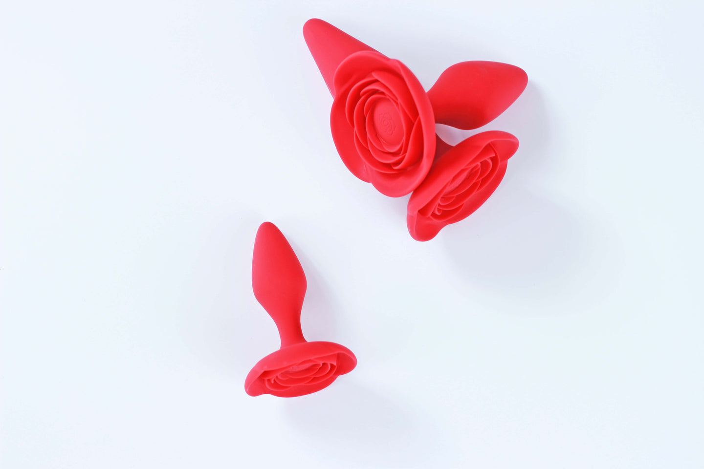 Small red rose butt plug laid in front of medium and large sizes of the same butt plug on white background