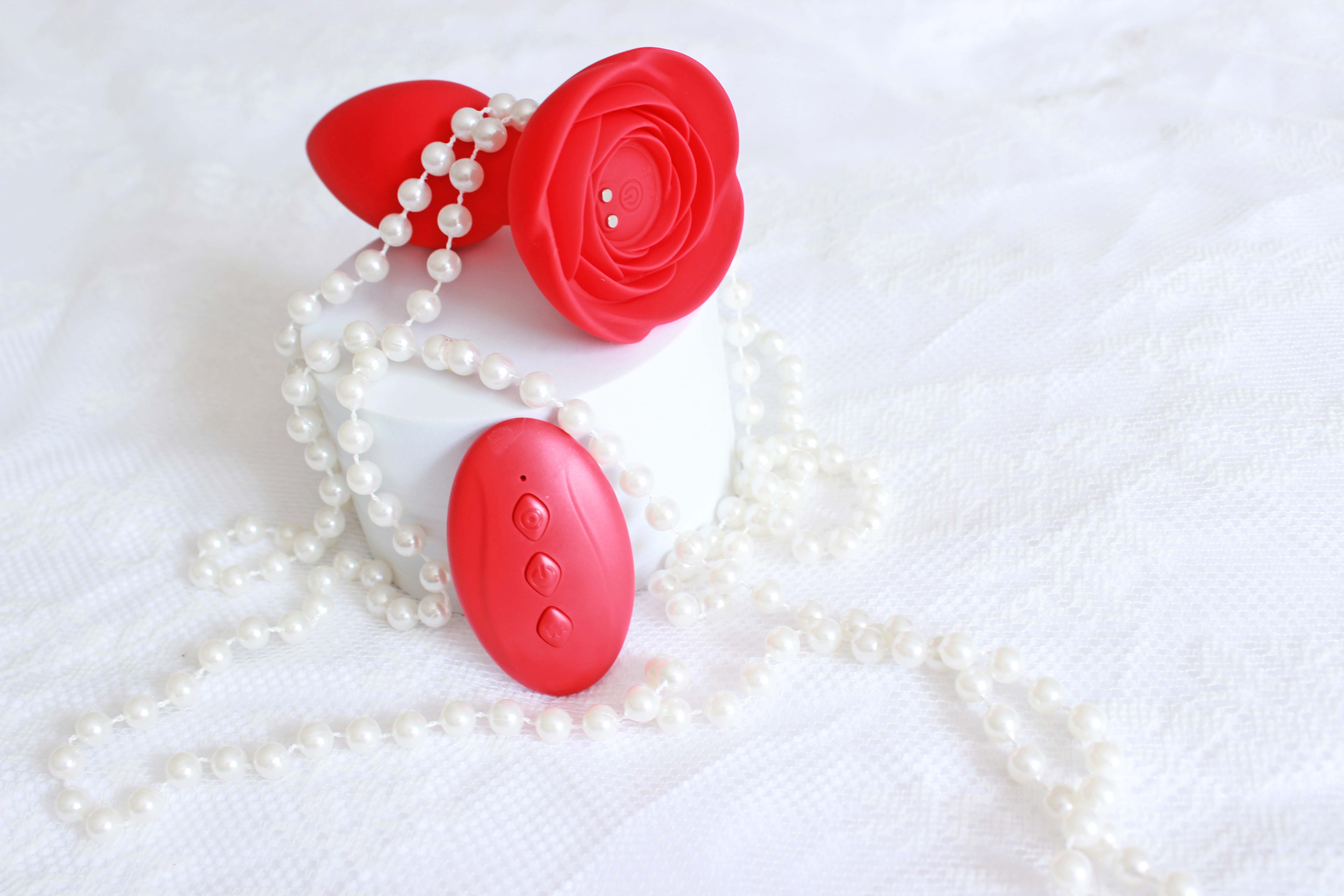 Red vibrating butt plug with rose base and remote control on a white lace background with pearls