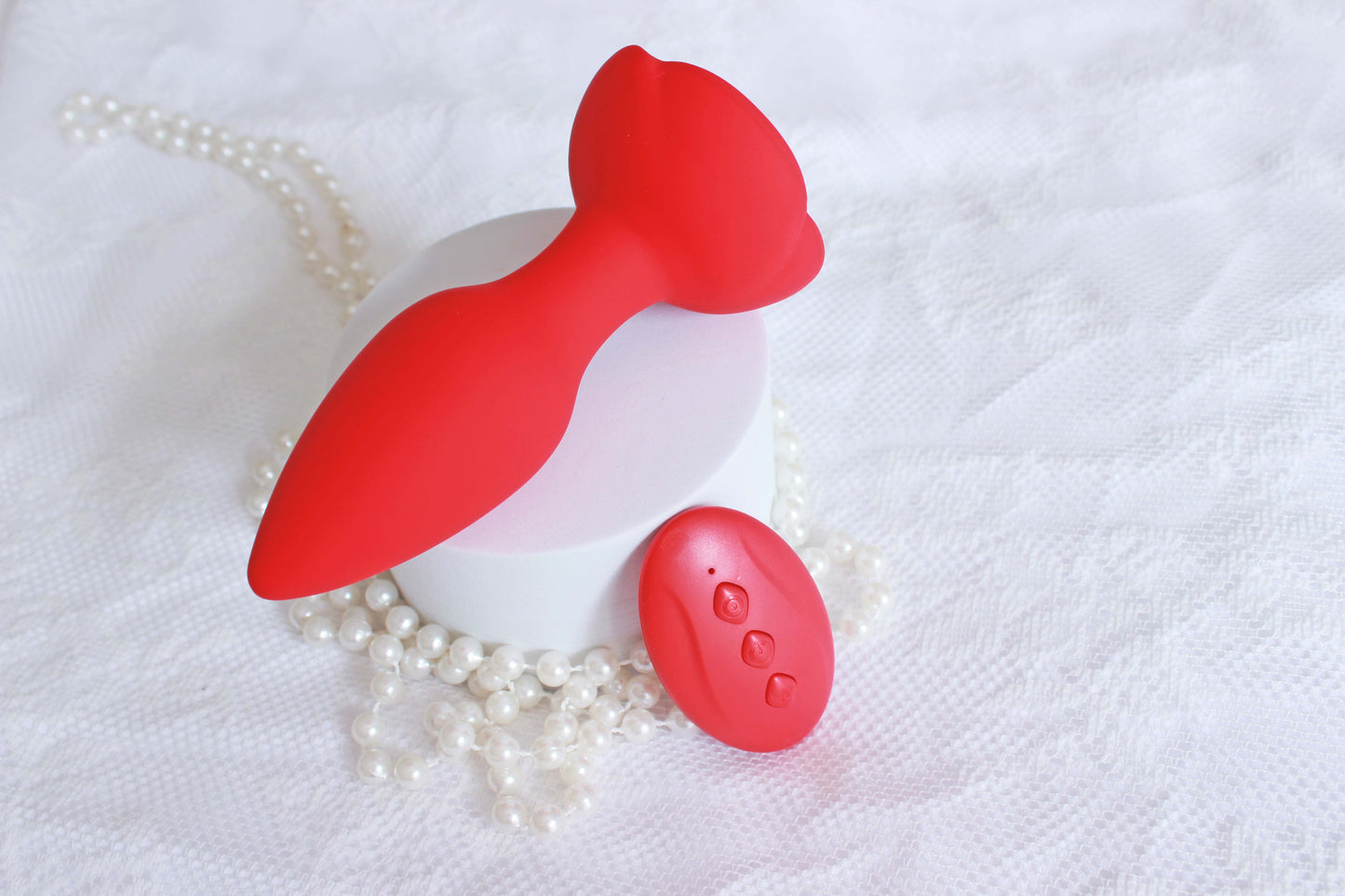 Down shot of red vibrating butt plug with rose base and remote control on a white lace background with pearls