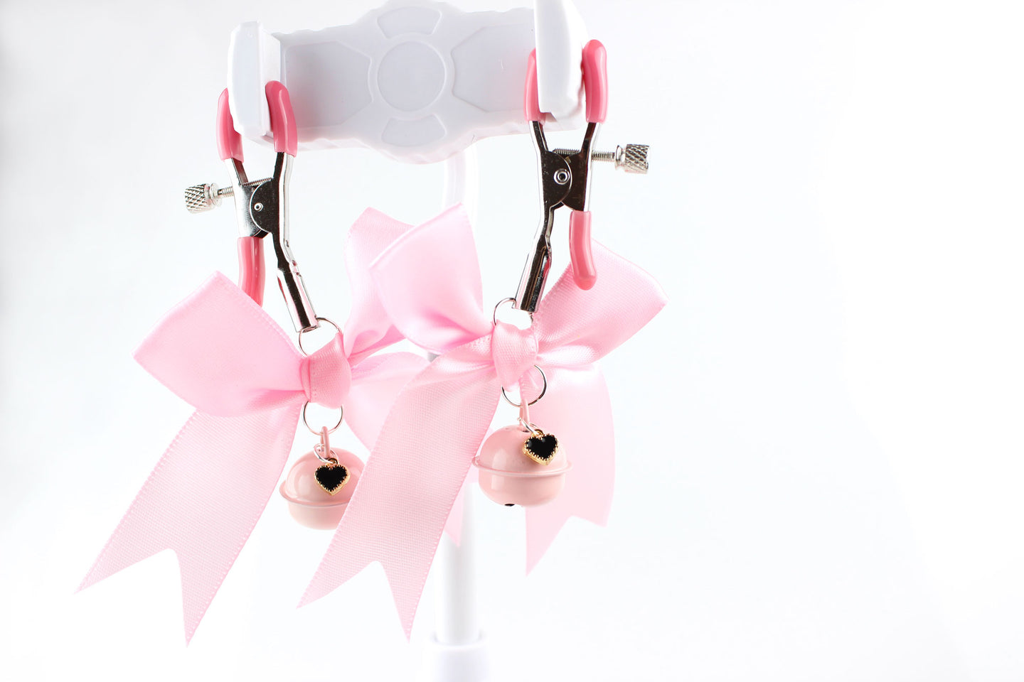 Two metal adjustable screw nipple clamps with pink PVC caps, pink satin bows, pink bells, and black heart charms hanging in front of a white background