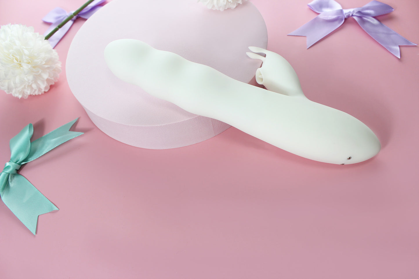 Back view of white bunny thruster dildo on pink background surrounded by purple and green bows and white flowers