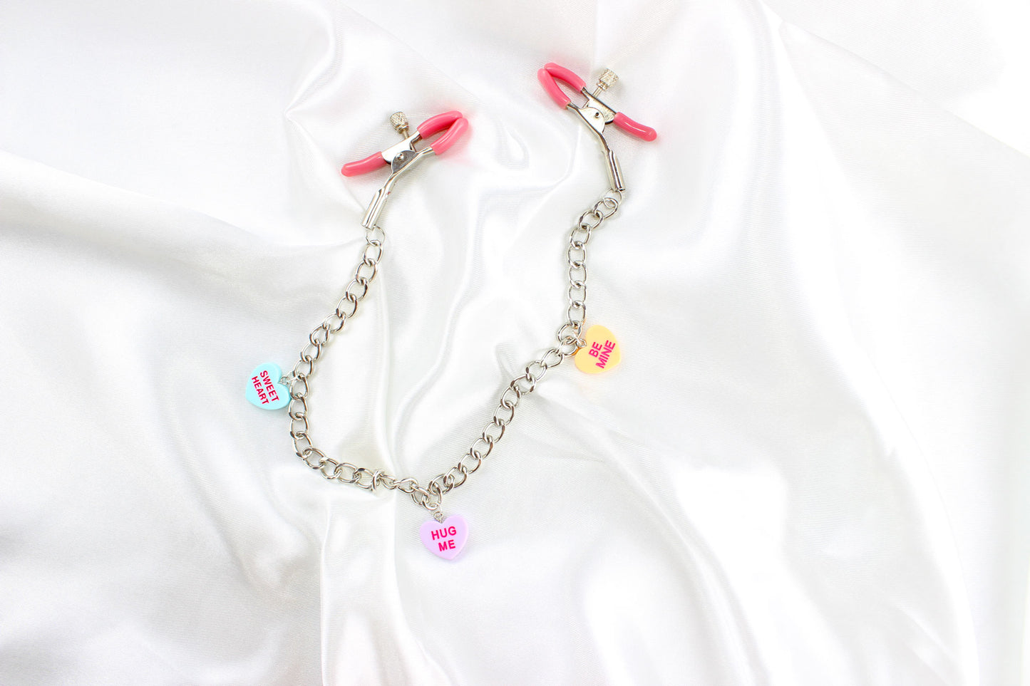 Pink PVC Capped silver tension screw nipple clamps connected by a chain with three candy heart charms on them placed on white satin fabric.