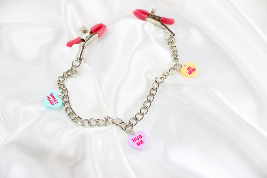 Pink PVC Capped silver tension screw nipple clamps connected by a chain with three candy heart charms on them placed on white satin fabric.