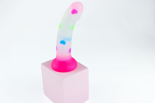 Opaque dildo with multicolored hearts and hot pink base on a pink block in front of a white background