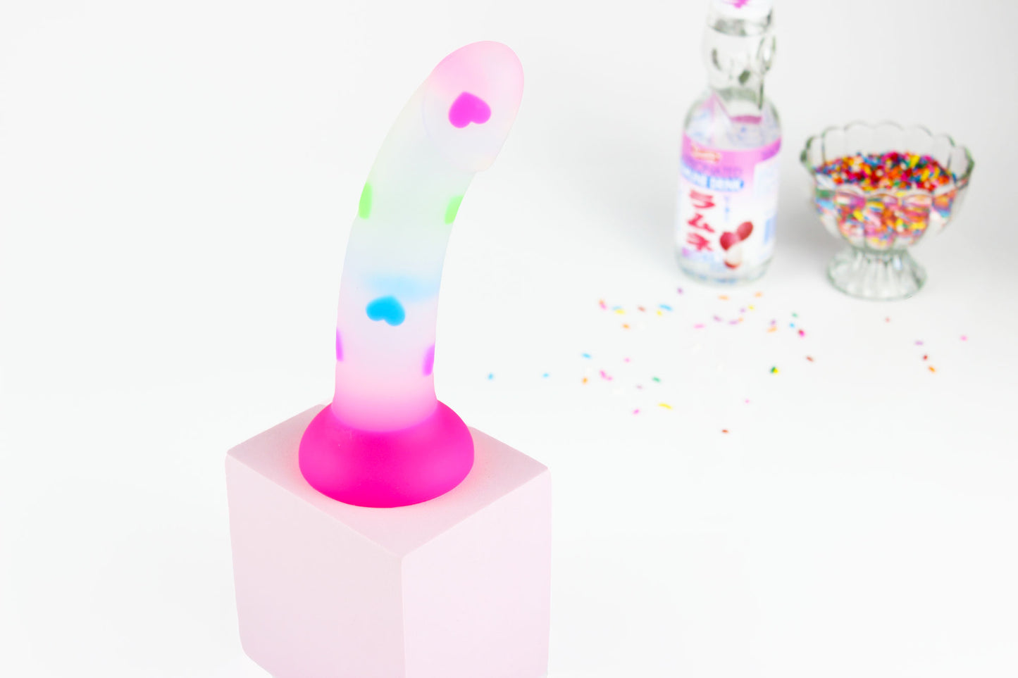 Opaque dildo with multicolored hearts and hot pink base on a pink block with out of focus soda and sprinkles in background