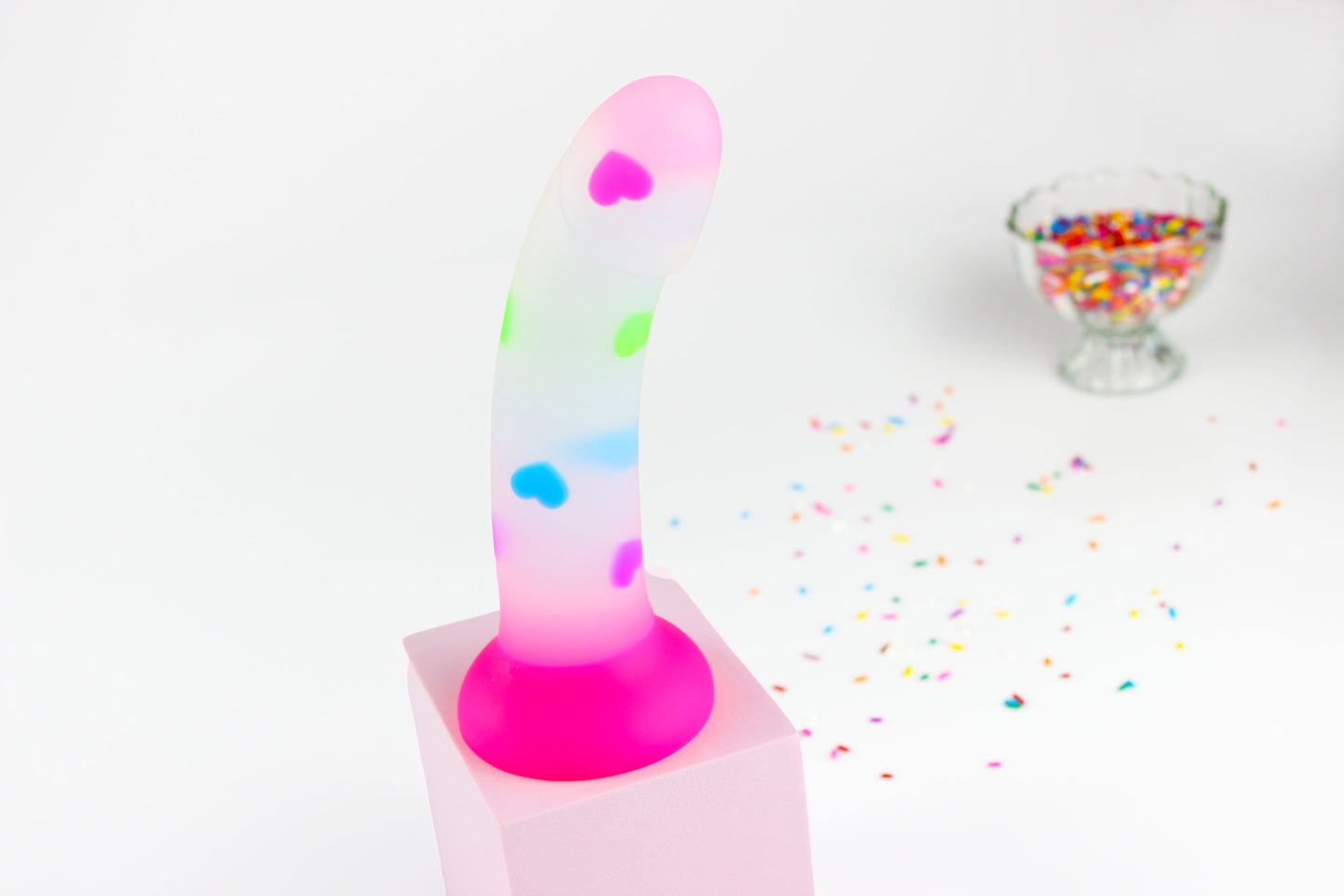 Opaque dildo with multicolored hearts and hot pink base on a pink block with out of focus sprinkles in background