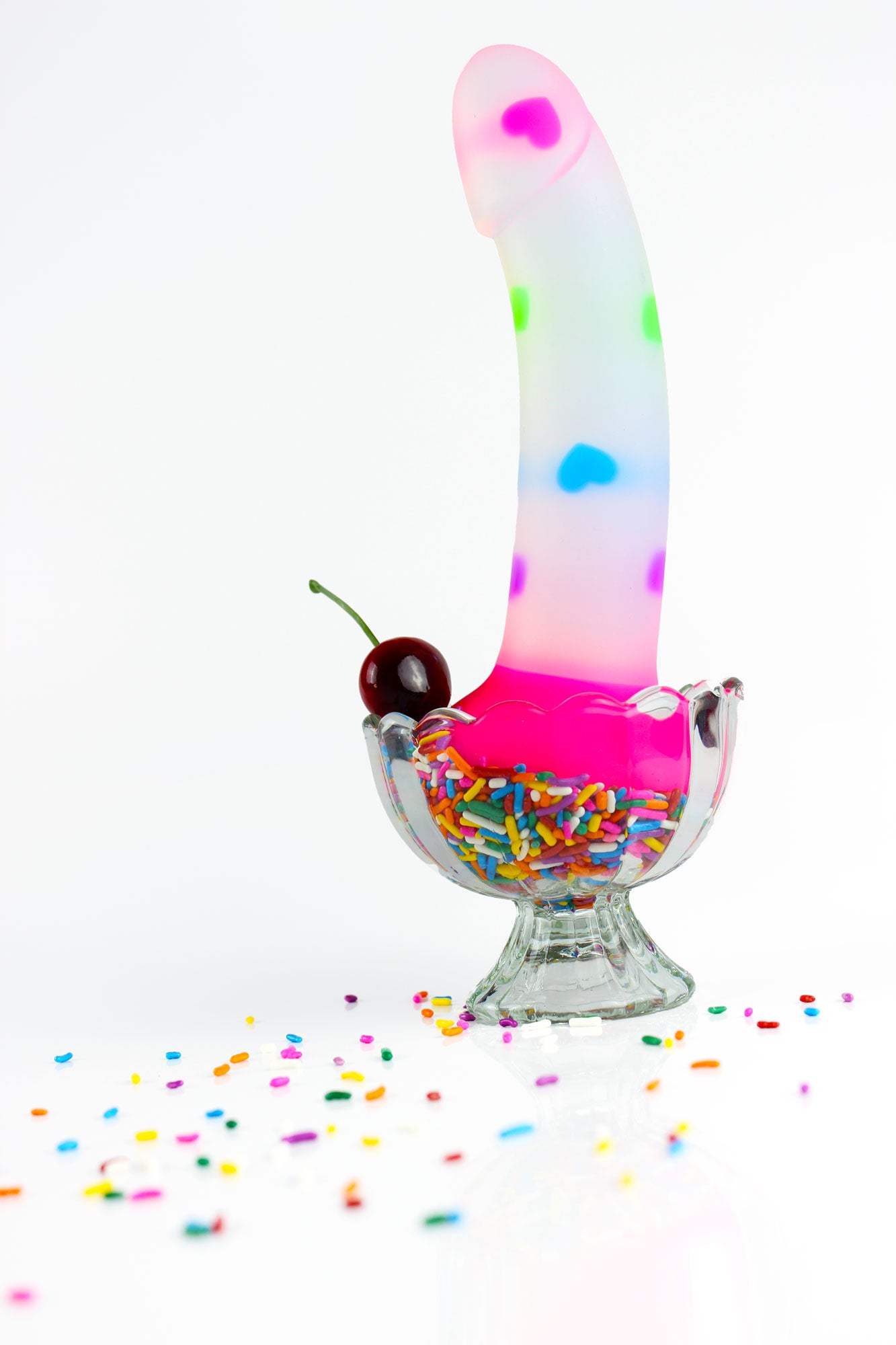 Opaque dildo with multicolored hearts and hot pink base in a glass dish of sprinkles with a cherry