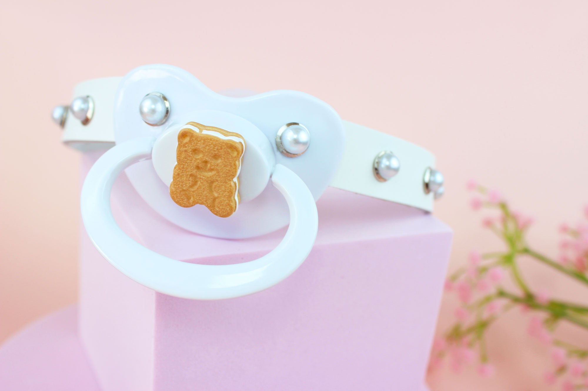 White adult pacifier gag with cookie bear charm and silver and pearl hardware on a pink stand against a pink background.
