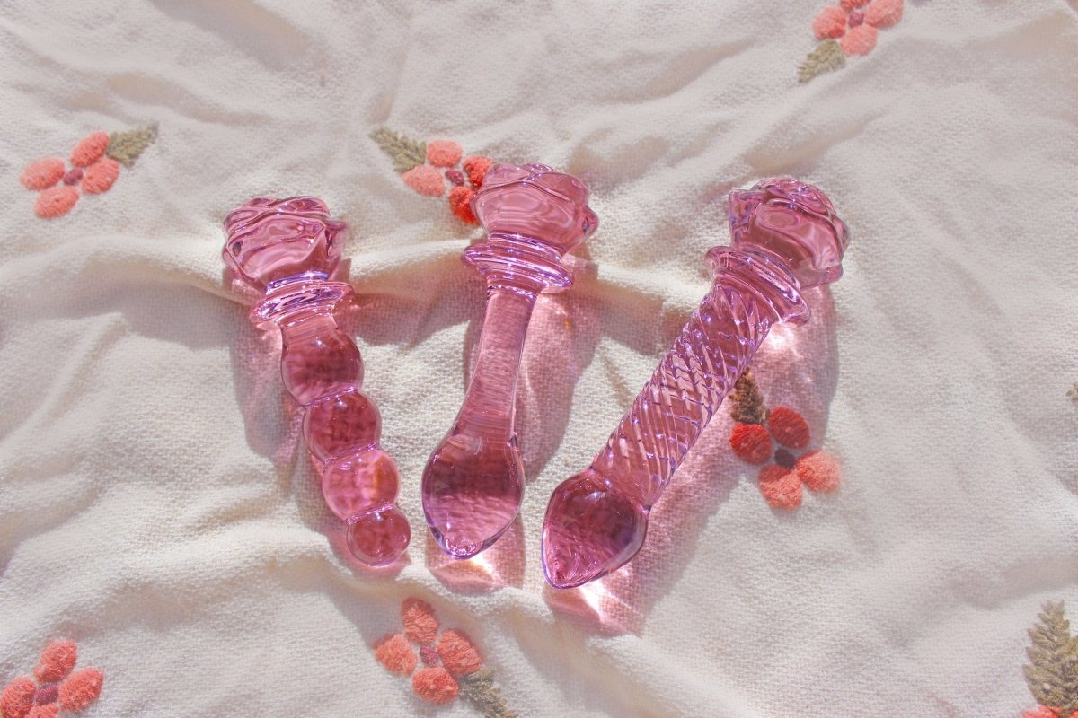 Three pink glass rose dildos laid out on a blanket. The far left has a pink rose dildo at the base with a knotted shaft. Middle dildo has glass rose base and butt plug shaft. Far right is a dildo with a rose base and a ridged shaft.
