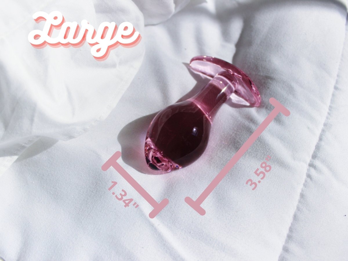 Large pink glass t bar butt plug against a white blanket with measurements reading 3.58 inches long by 1.34 inches wide.