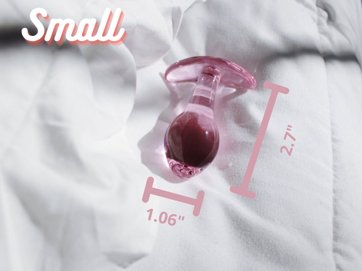 Small pink glass t bar butt plug laid out on a blanket with measurements of 2.7 inches tall by 1.06 inches wide at the base.