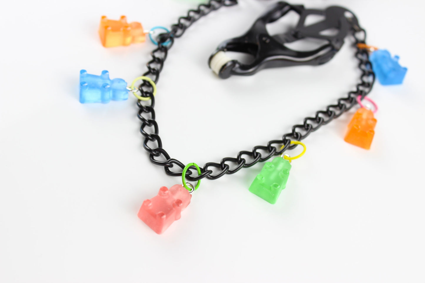 Close up image of Black Japanese clover clamps with a black chain connecting the clamps and 6 colorful gummy bear charms hanging off the chain placed on a white background.