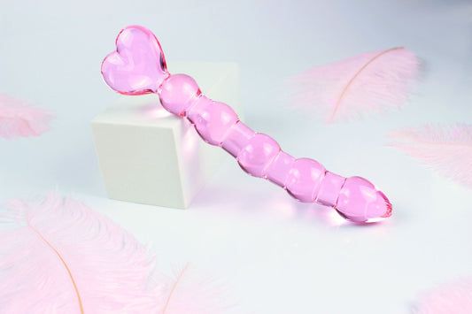 Pink glass anal bead dildo with heart shaped base on white background with pink feathers