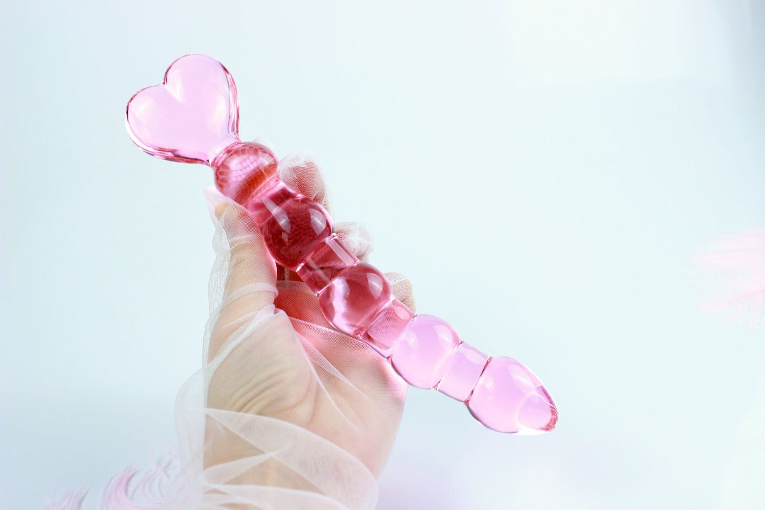 Gloved hand holding pink glass anal bead dildo with heart shaped base over white background