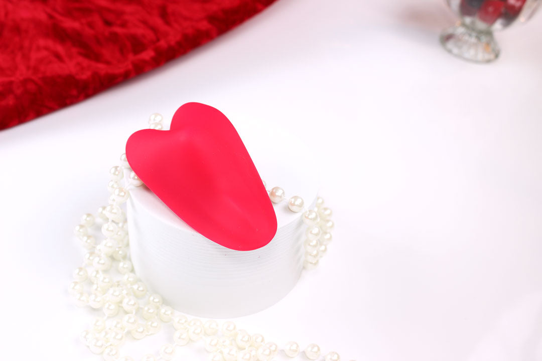 Red heart shaped silicone remote controlled panty vibrator laid out on a white stand with red velvet cloth, cherries, and pearls around it.