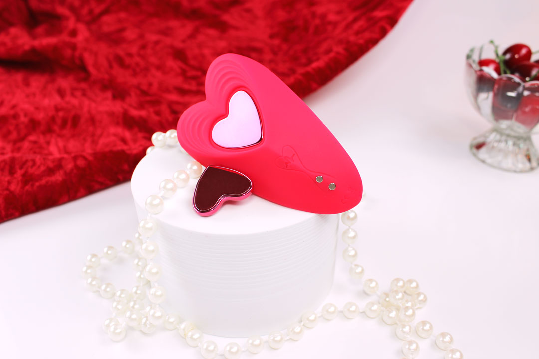 Red heart shaped silicone remote controlled panty vibrator laid out on a white stand with red velvet cloth, cherries, and pearls around it.