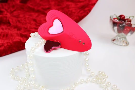 Red heart shaped silicone remote controlled panty vibrator  laid out on a white stand with red velvet cloth, cherries, and pearls around it.