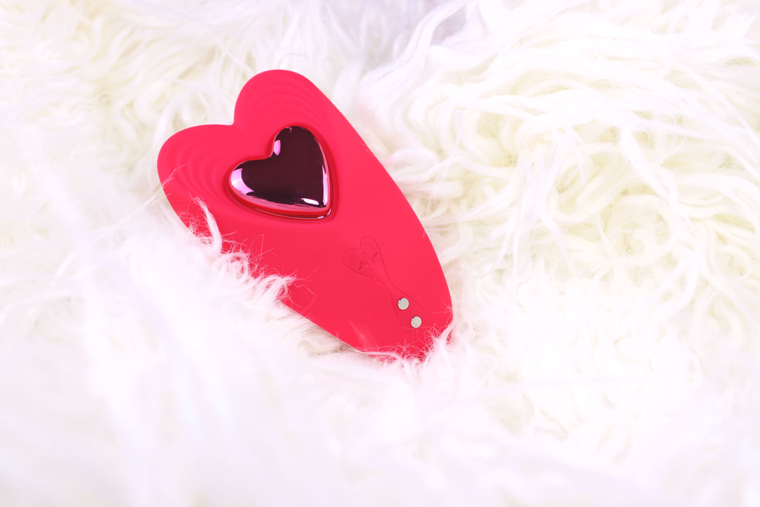 Red heart shaped silicone panty vibrator and remote laying in a fluffy white blanket.