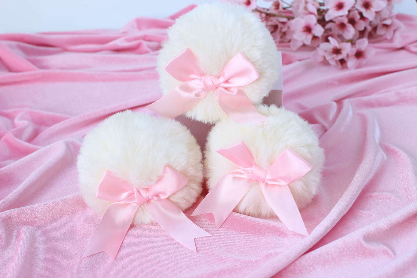 Set of three cute white bunny tail glass butt plugs with pink bows on the pom against a pink background with pink flowers.