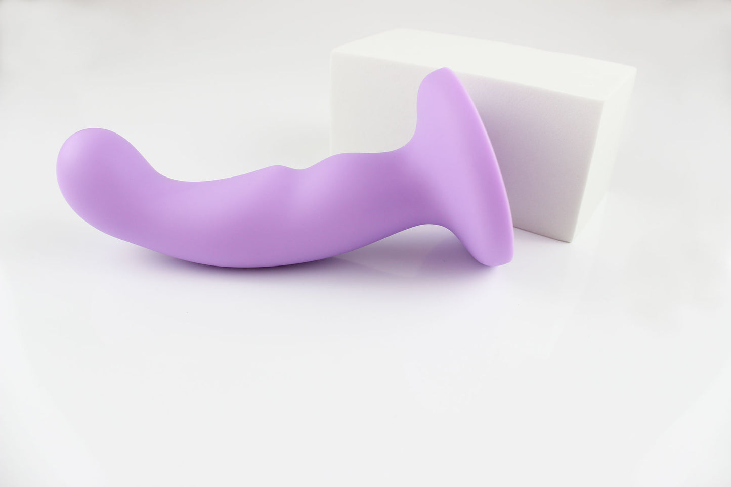 Light purple silicone dildo laying sideways in front of white rectangular block on white background