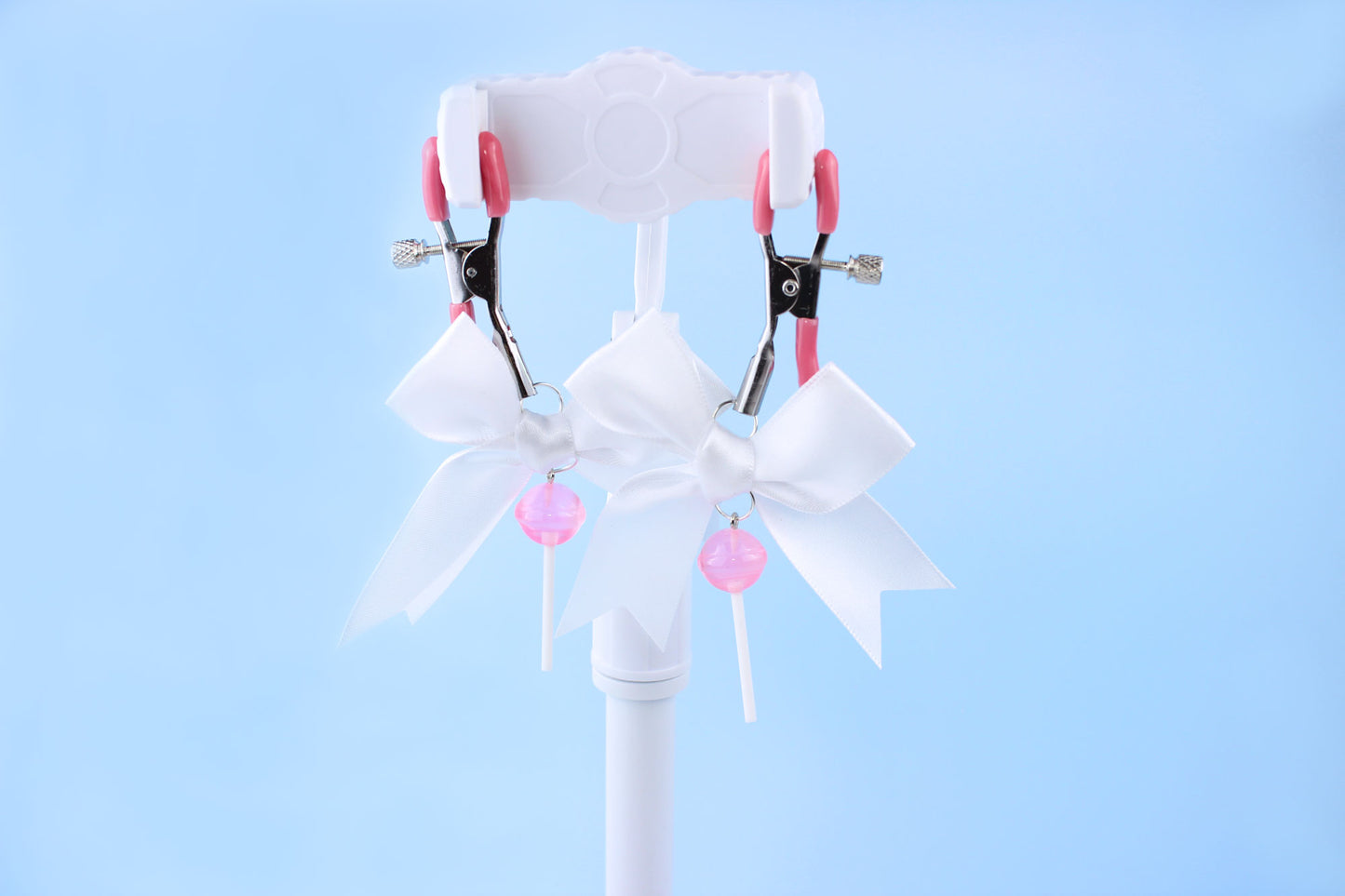 Pink grip silver screw nipple clamps with a white bow and a pink lollipop hanging off of each clamp in front of a blue background.