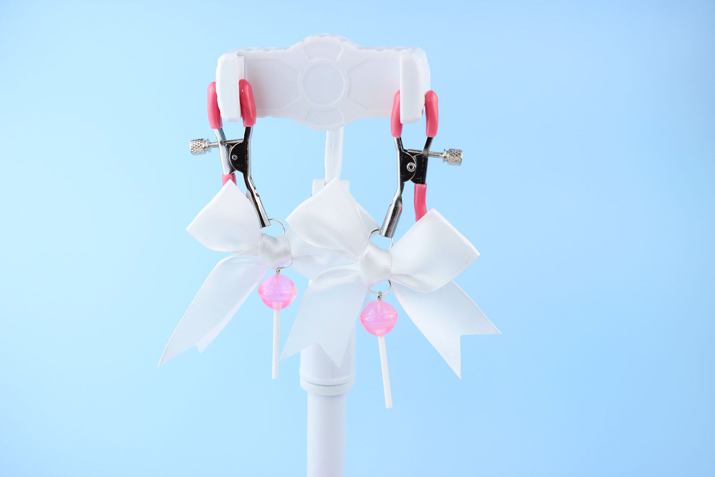 Pink grip silver screw nipple clamps with a white bow and a pink lollipop hanging off of each clamp in front of a blue background.