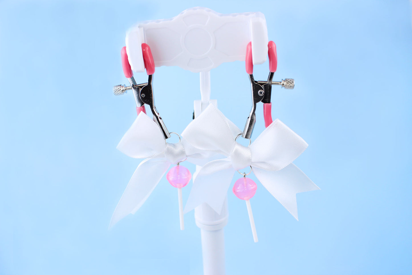 Pink grip silver screw nipple clamps with a white bow and a pink lollipop hanging off of each clamp in front of a blue background.