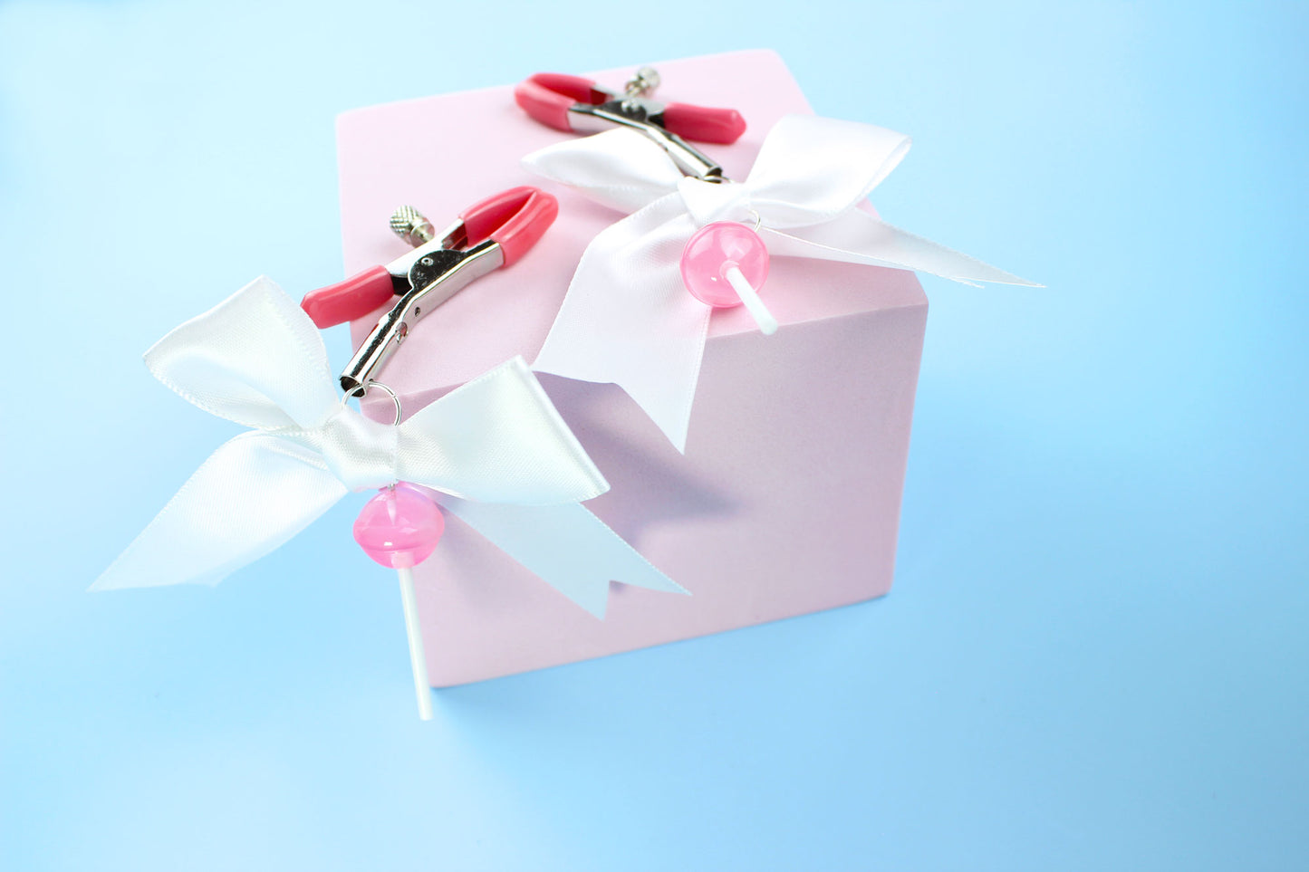 Pink grip silver screw nipple clamps with a white bow and a pink lollipop hanging off of each clamp on a pink square over blue background.