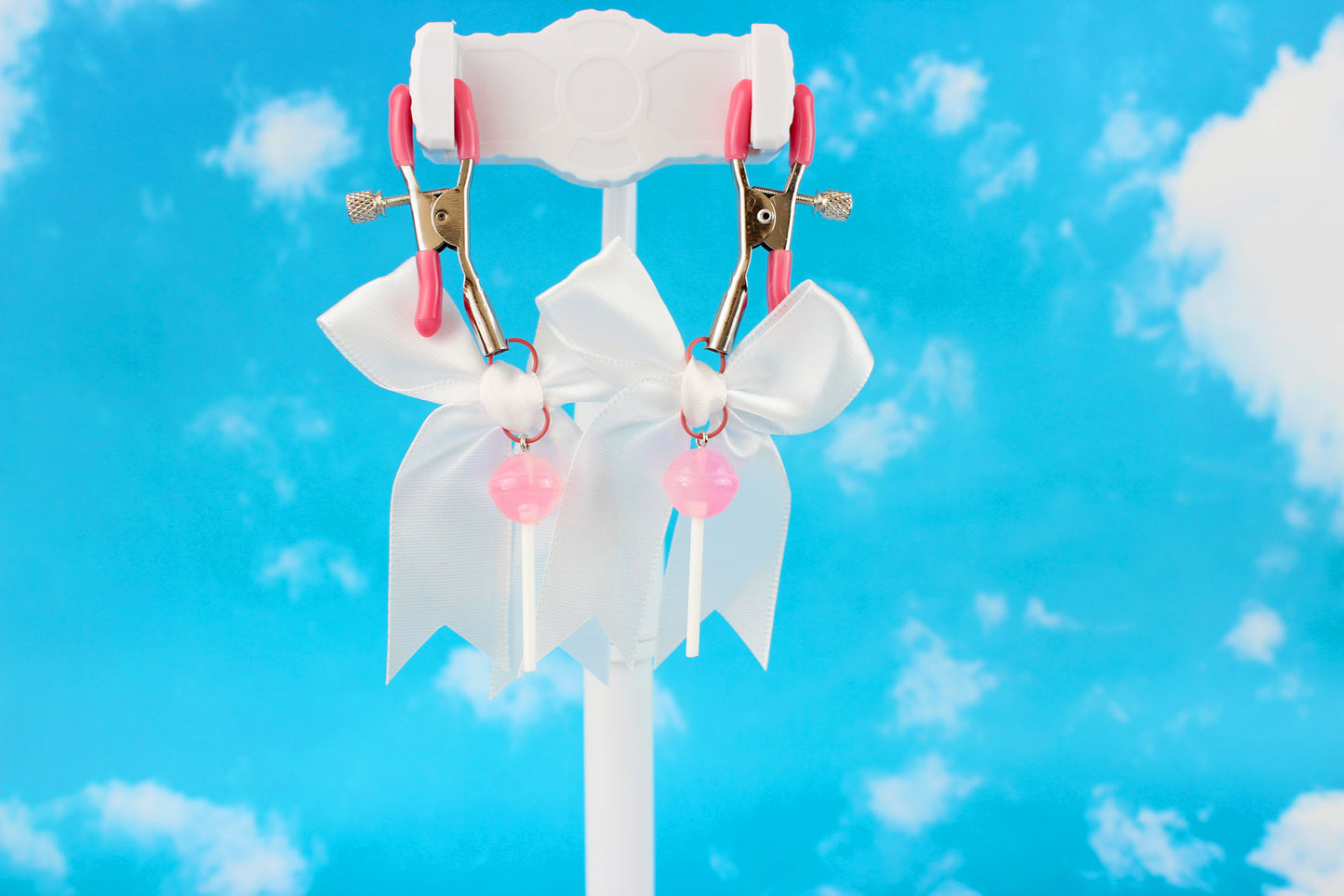 Pink grip silver screw nipple clamps with a white bow and a pink lollipop hanging off of each clamp in front of a sky background.