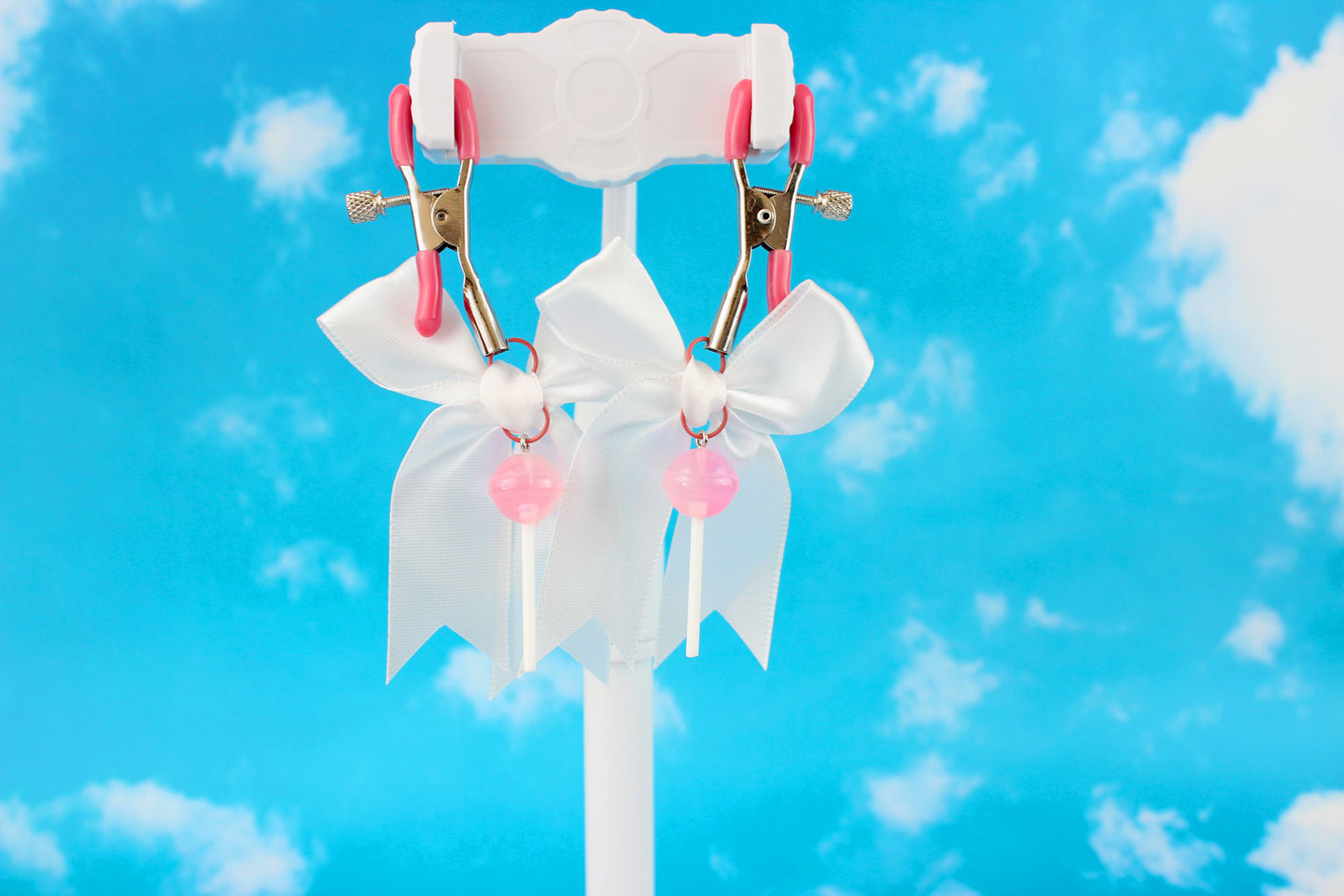 Pink grip silver screw nipple clamps with a white bow and a pink lollipop hanging off of each clamp in front of a sky background.