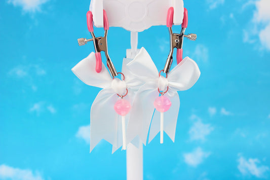 Pink grip silver screw nipple clamps with a white bow and a pink lollipop hanging off of each clamp in front of a sky background.