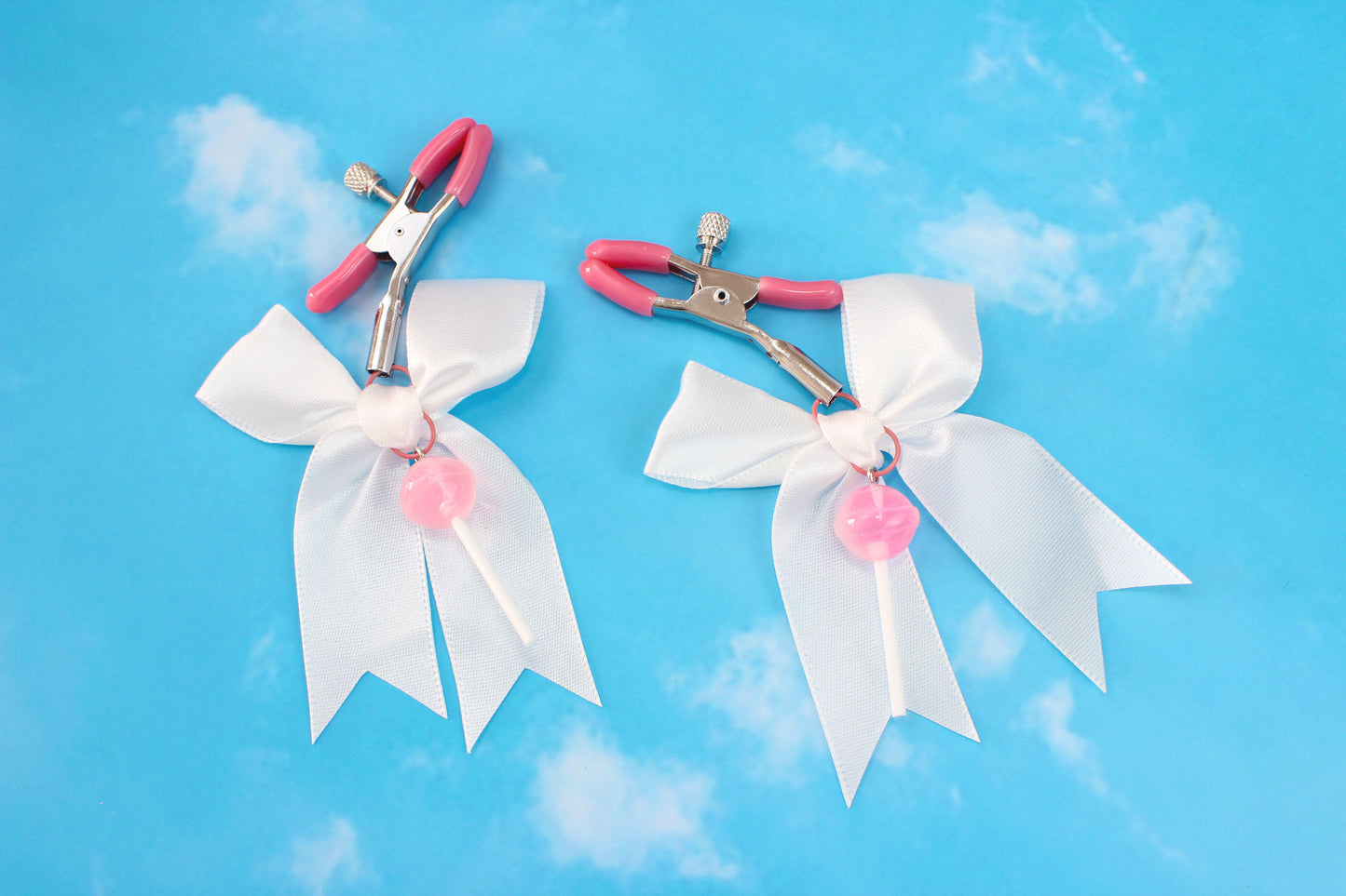 Pink grip silver screw nipple clamps with a white bow and a pink lollipop hanging off of each clamp in front of a sky background.