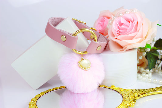 Pink leather collar with gold hardware, pink pom charm, and gold pearl heart charm surrounded by roses and a hand mirror