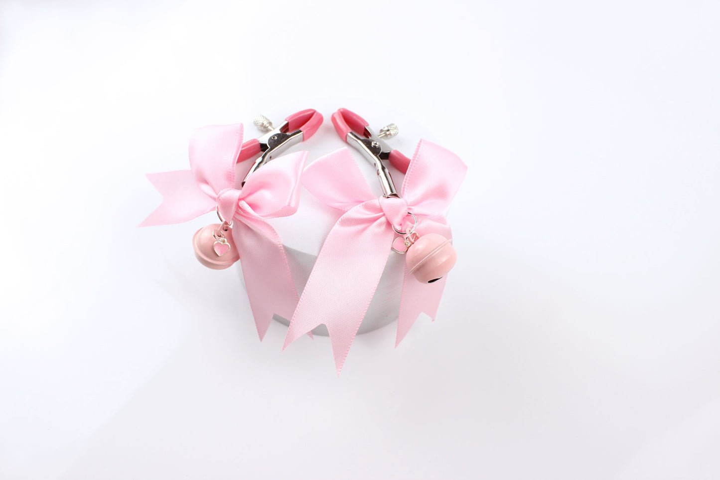 Two metal adjustable screw nipple clamps with pink PVC caps, pink satin bows, pink bells, and pink heart charms laying over a foam circle on a white background