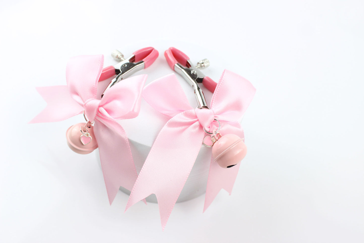 Two metal adjustable screw nipple clamps with pink PVC caps, pink satin bows, pink bells, and pink heart charms laying over a foam circle on a white background